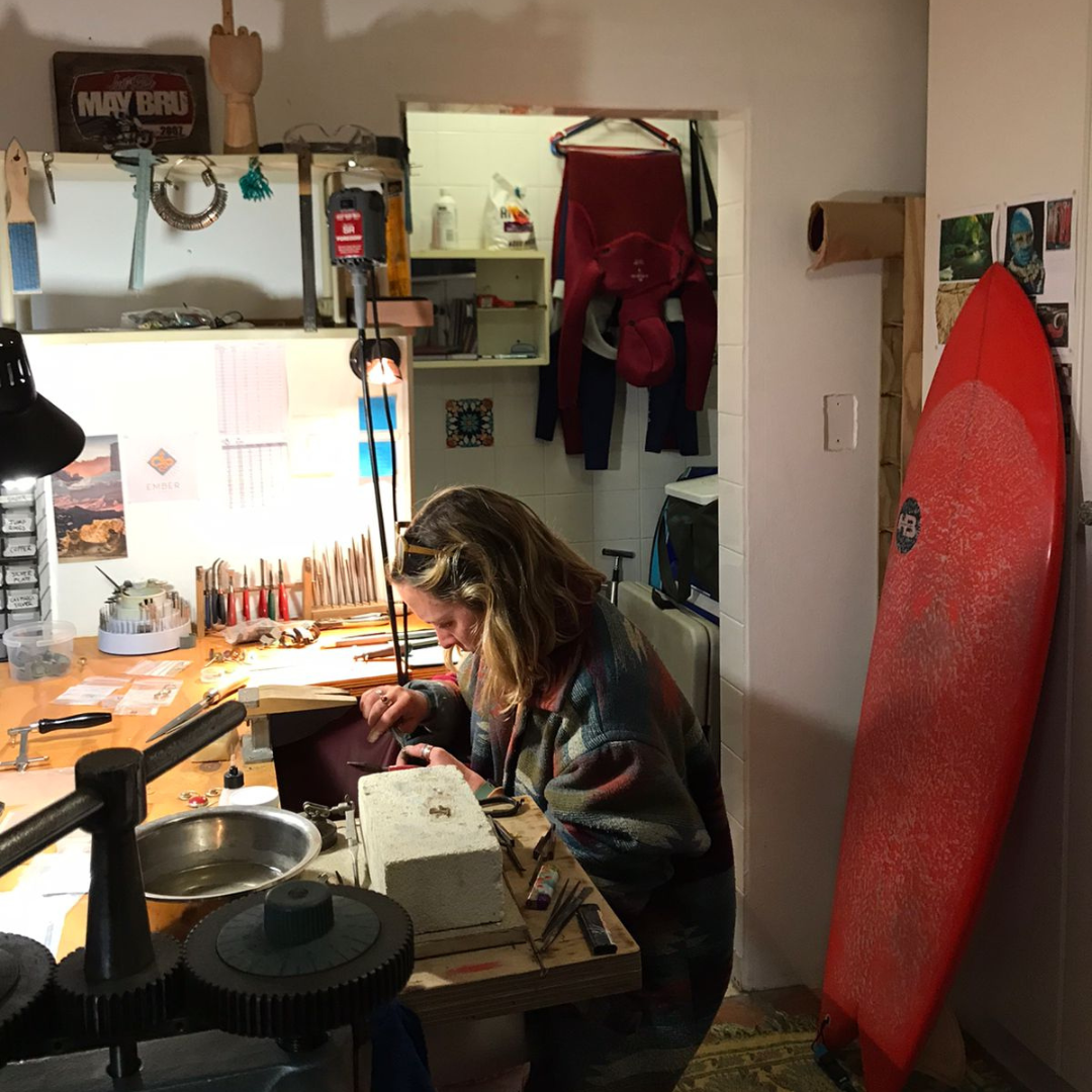 Person working at a desk with tools and supplies, red surfboard leaning against a wall.