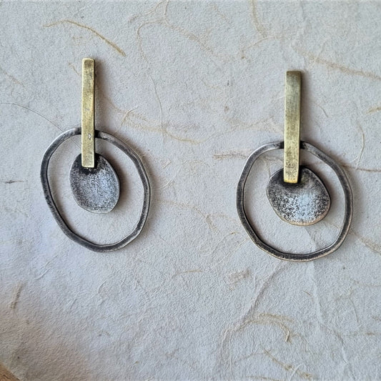 A pair of handmade dangle earrings featuring textured silver-toned rings, a central hammered silver disc, and a vertical gold-toned post, displayed on a textured off-white surface.
