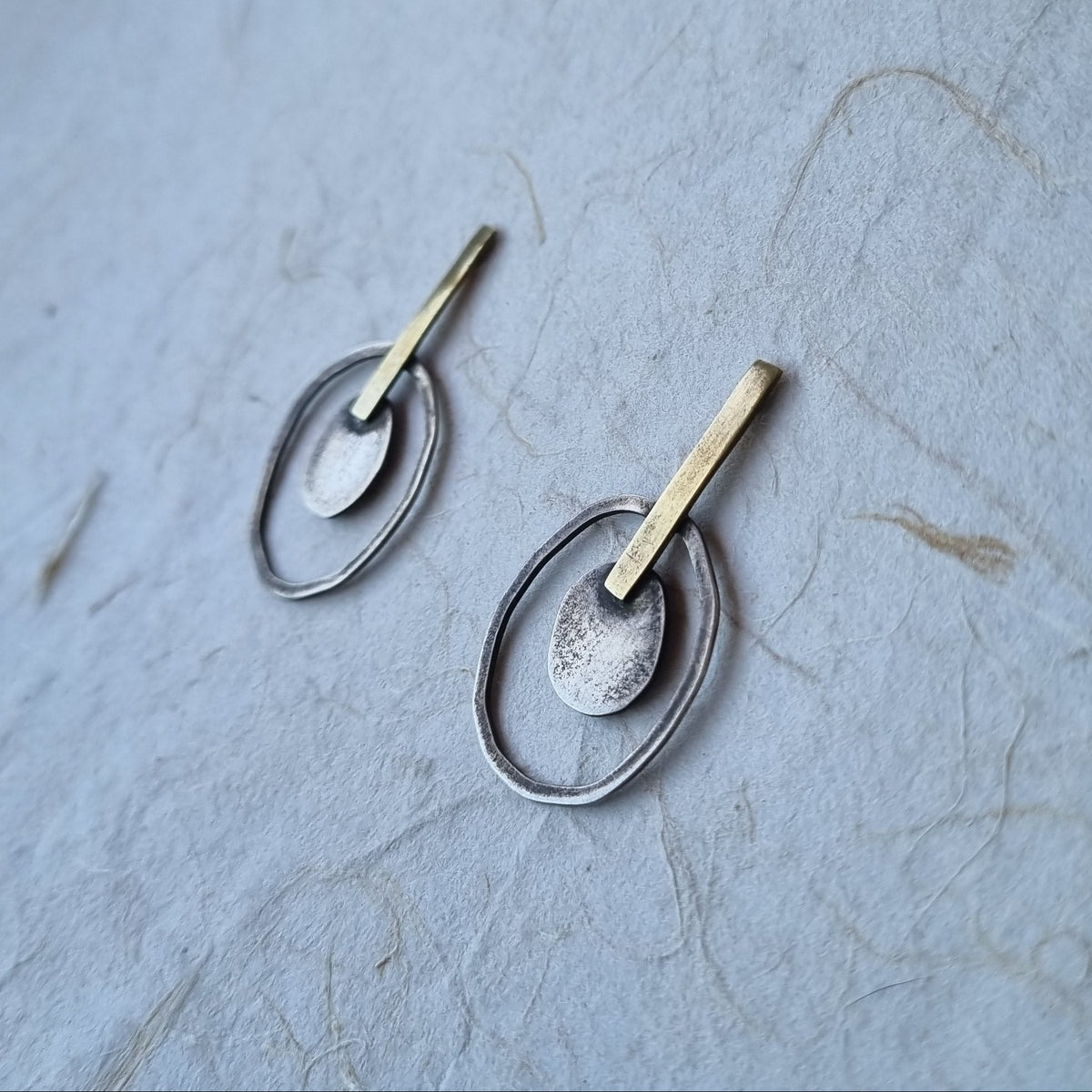 A pair of handmade dangle earrings with textured silver and gold metal, displayed on a textured light blue paper background.