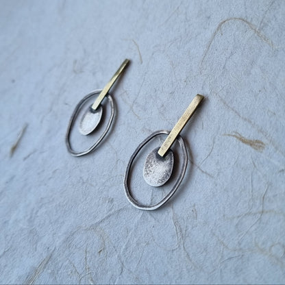 A pair of handmade dangle earrings with textured silver and gold metal, displayed on a textured light blue paper background.