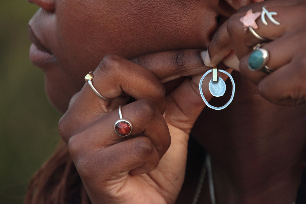 Close-up of a person wearing a silver hoop earring with a circular disc pendant, while wearing other rings on their fingers.