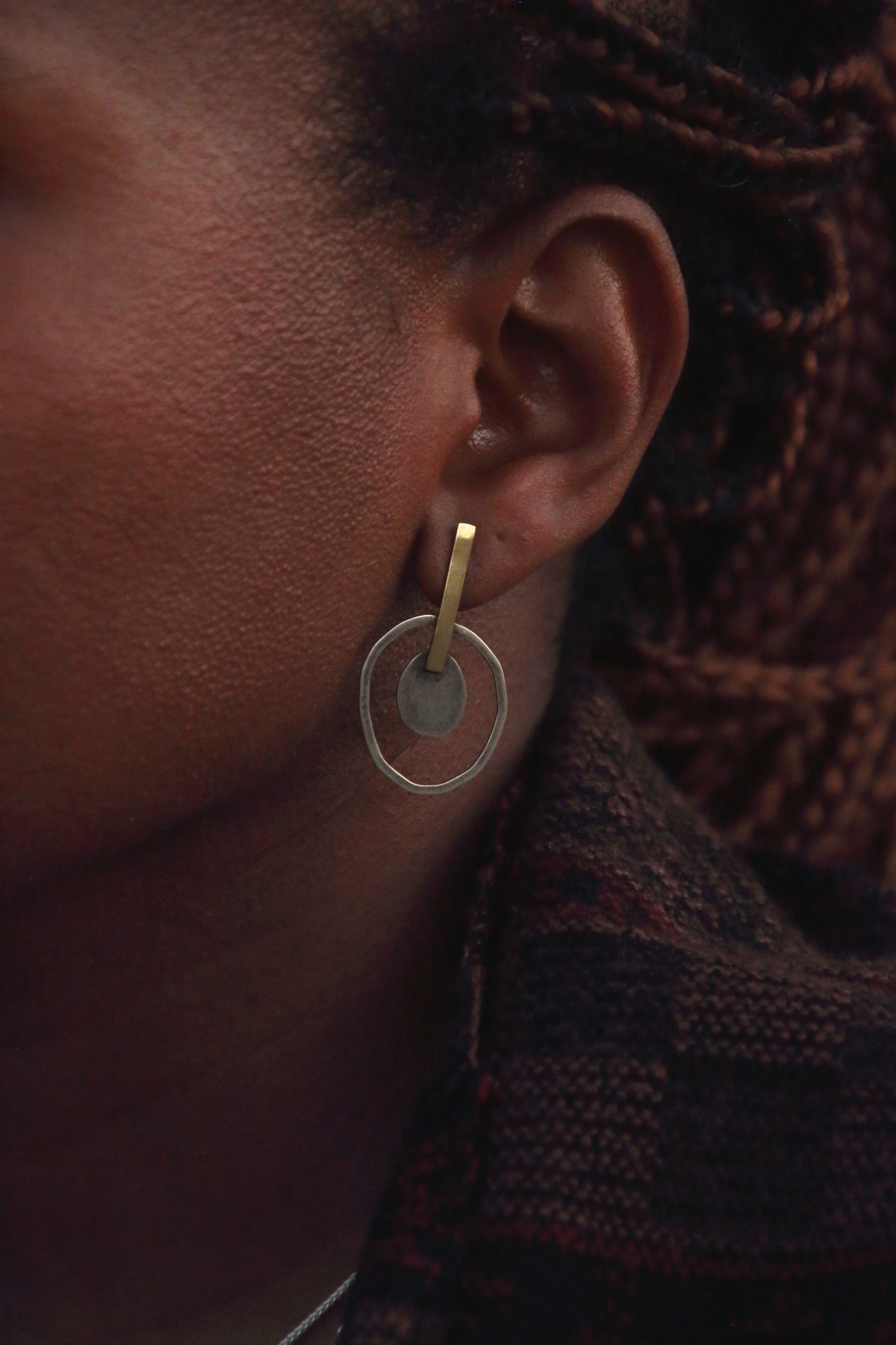 A close-up of a person's ear wearing a minimalist gold hoop earring with a circular disc detail, set against dark braided hair and a textured dark knit garment.