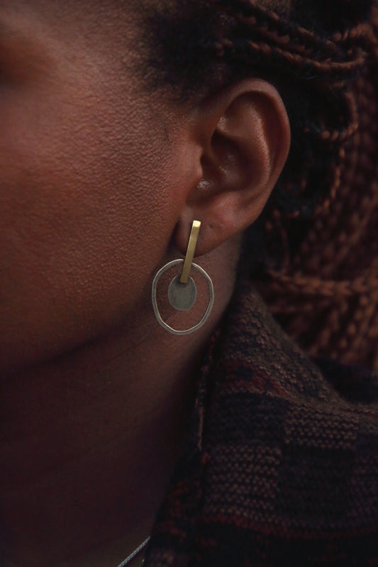 A close-up of a person's ear wearing a minimalist gold hoop earring with a circular disc detail, set against dark braided hair and a textured dark knit garment.