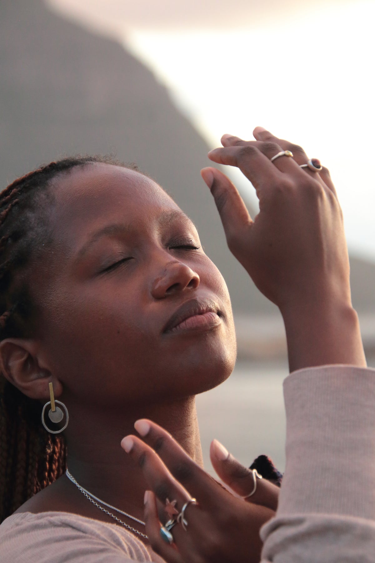 A close-up lifestyle shot of a woman with her eyes closed, wearing gold hoop earrings, a silver chain necklace, and multiple rings, with her hands raised in a serene pose against a soft-focus outdoor background.