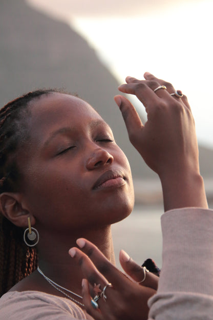 A close-up lifestyle shot of a woman with her eyes closed, wearing gold hoop earrings, a silver chain necklace, and multiple rings, with her hands raised in a serene pose against a soft-focus outdoor background.